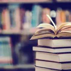 Otwarta księga na stosie książek, jakby opowiadała historie o lalka motywy. W tle regały pełne literatury.