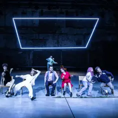 Scena teatralna z grupą ludzi w różnych pozach, jakby w trakcie pantomimy. W tle neonowy prostokąt.