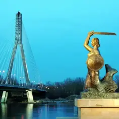 Syrenka Warszawska z mieczem i tarczą, na tle nowoczesnego mostu. Symbol miasta, herb Warszawy, czuwa nad Wisłą.