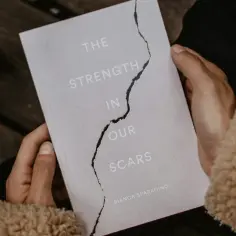 Dłonie trzymają książkę "The Strength in Our Scars", która opowiada o przezwyciężaniu cierpienia. Obok kawa z pianką.