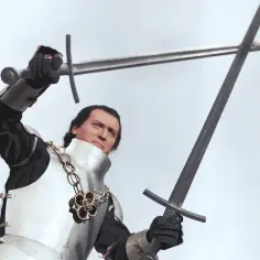 Rycerz w zbroi, z dwoma mieczami w dłoniach, gotowy do walki. W tle widać krzyżaków.