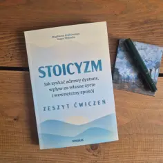 Zeszyt ćwiczeń "Stoicyzm co to" na drewnianym blacie. Książka pomaga zyskać dystans i spokój. Obok zielone pióro.