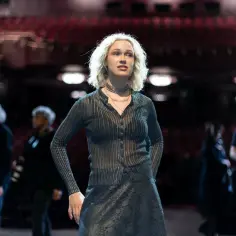 Ofelia, młoda kobieta o blond włosach, stoi na scenie teatru, ubrana w błyszczącą bluzkę i spódnicę.