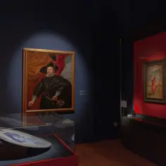 Portret mężczyzny w kapeluszu i bogatym stroju, z rubensowskimi kształtami w tle. Obok w gablocie ceramiczna taca.