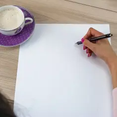 Dłoń z długopisem nad białą kartką, obok filiżanka kawy na fioletowym talerzyku.