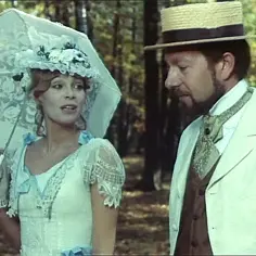 Kobieta w białej sukni i kapeluszu z woalką, z parasolką, obok mężczyzny w słomkowym kapeluszu i jasnej marynarce. Scena jak z filmu "Praca u podstaw", gdzie lalka może symbolizować sztuczność.