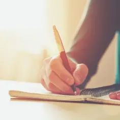 Dłoń pisze piórem w zeszycie, tworząc peryfrazę myśli.