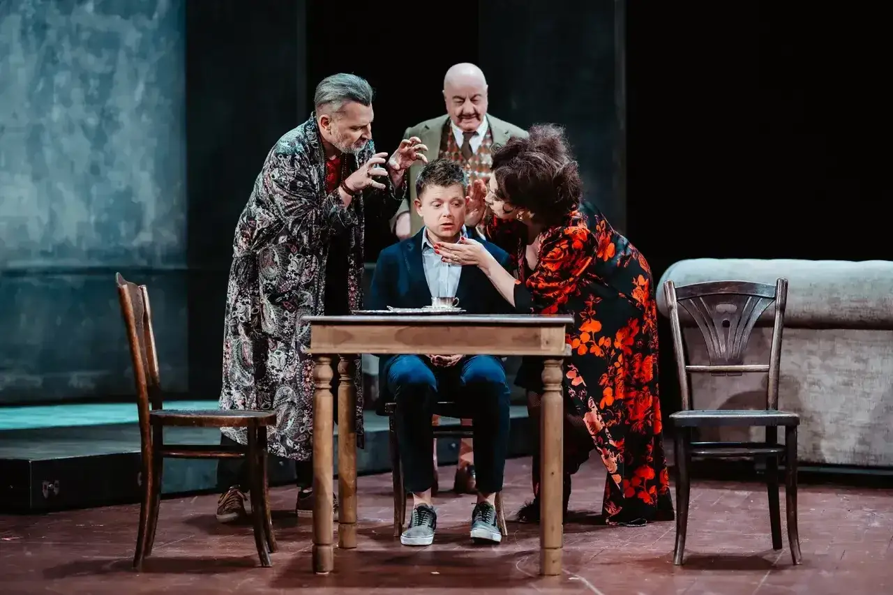 Tango bohaterowie w dramatycznej scenie. Mężczyzna siedzi przy stole, otoczony przez trzy postacie, które zdają się go naciskać.